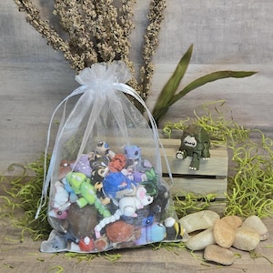 May include: A white organza drawstring bag overflowing with colourful miniature figurines. A small green and brown toy creature sits on a wooden crate. Several small, light-coloured stones are scattered nearby, with dried plants in the background.