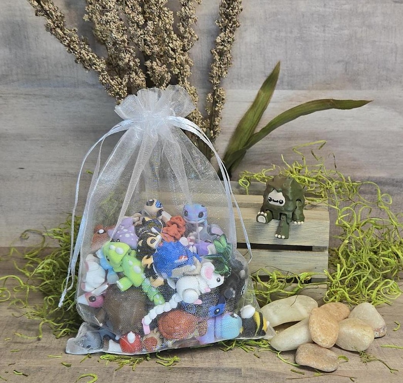 May include: A white organza drawstring bag overflowing with colourful miniature figurines. A small green and brown toy creature sits on a wooden crate. Several small, light-coloured stones are scattered nearby, with dried plants in the background.