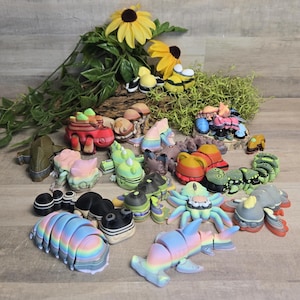 May include: A collection of colorful, whimsical figurines, including insects, sea creatures, and mushrooms. The figurines are in various colors, including blue, green, pink, and yellow. The arrangement includes a background of yellow flowers and greenery.