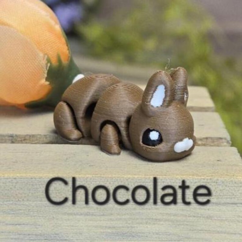 K&ouml;nnte beinhalten: Eine braune, segmentierte Hasenfigur mit wei&szlig;en Akzenten an Ohren und Gesicht. Das Wort "Chocolate" ist in Schwarz auf der Holzoberfl&auml;che darunter gedruckt. Der Hase befindet sich auf einer Holzoberfl&auml;che.