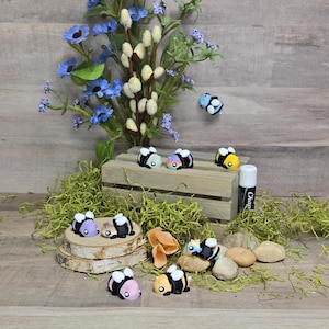 Puede incluir: Una colección de figuritas de abejas pintadas a mano en varios colores, incluyendo negro, blanco, amarillo, rosa y azul. Las figuritas están dispuestas en una caja de madera y una rodaja de tronco de abedul. Las abejas están rodeadas de musgo verde y pequeñas piedras.