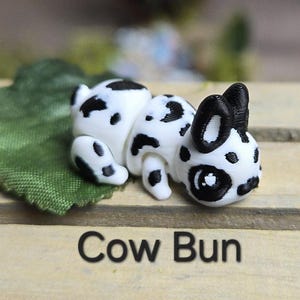 K&ouml;nnte beinhalten: Eine wei&szlig;-schwarz gefleckte Figur in Form eines Kuhbr&ouml;tchens, die auf einem gr&uuml;nen Blatt liegt. Der Text "Cow Bun" ist unten im Bild in Schwarz gedruckt. Die Figur hat schwarze Ohren und schwarze Flecken.