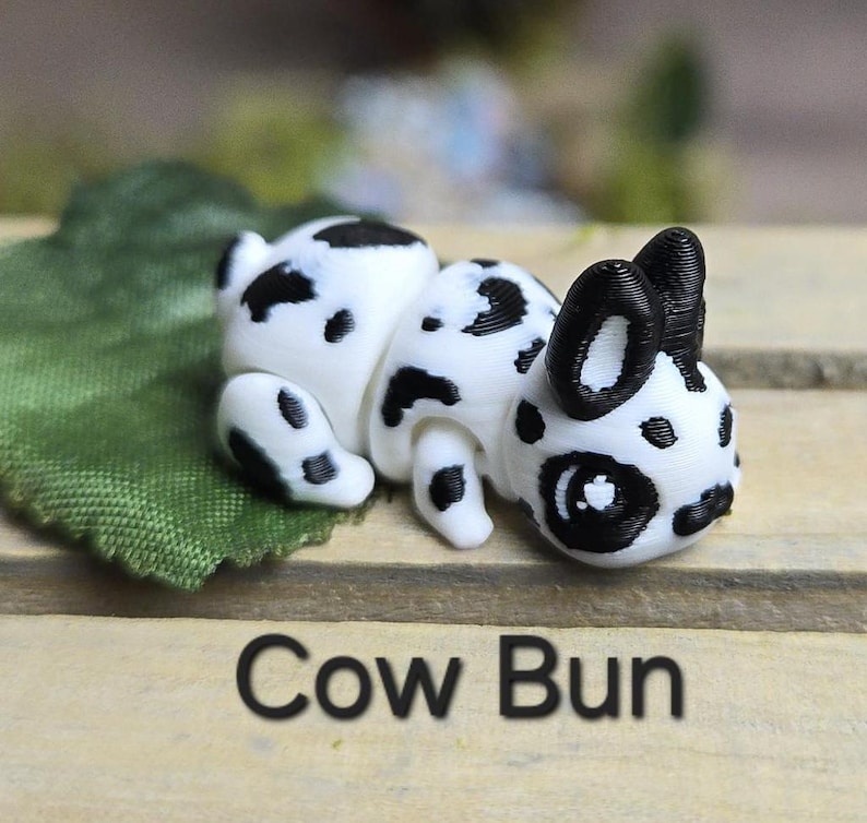 K&ouml;nnte beinhalten: Eine wei&szlig;-schwarz gefleckte Figur in Form eines Kuhbr&ouml;tchens, die auf einem gr&uuml;nen Blatt liegt. Der Text "Cow Bun" ist unten im Bild in Schwarz gedruckt. Die Figur hat schwarze Ohren und schwarze Flecken.