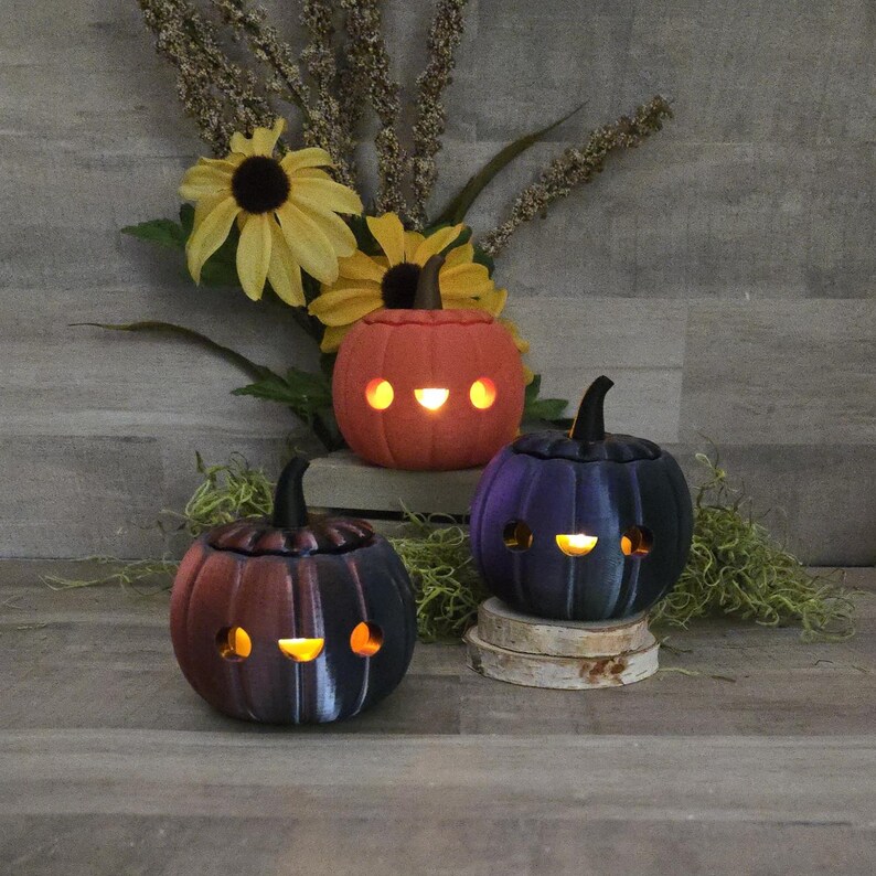 May include: Three ceramic pumpkin lanterns in orange, purple, and black, each with a carved face and a small stem. The pumpkins are lit from within, creating a warm glow. They are displayed on a wooden surface with sunflowers and greenery.