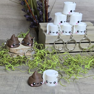 May include: A set of keychains featuring cute cartoon faces. There are two brown poop keychains and three white toilet paper roll keychains. Each keychain has a silver ring and a silver chain.
