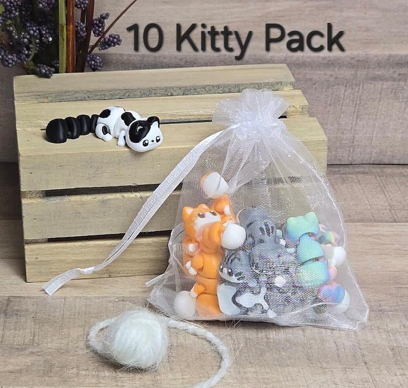 K&ouml;nnte beinhalten: Eine wei&szlig;e Organzabtasche gef&uuml;llt mit 10 kleinen, bunten Katzenfiguren. Die Tasche ist mit einem wei&szlig;en Band gebunden. Der Text "10 Kitty Pack" ist &uuml;ber der Tasche sichtbar.