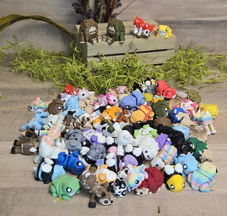 May include: A selection of colourful miniature animal figurines, including frogs, deer, and bears, in various poses and colours. Some figurines are on a wooden crate with greenery.