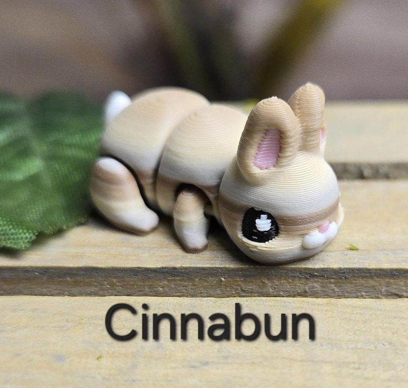 K&ouml;nnte beinhalten: Hellbraune Hasenfigur mit cremefarbenem K&ouml;rper und rosa Innenohren. Der Hase hat gro&szlig;e schwarze Augen und liegt. Das Wort "Cinnabun" ist unten im Bild in Schwarz gedruckt. Die Figur steht auf einer Holzoberfl&auml;che.