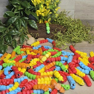 Puede incluir: Una colección de coloridos gusanos de juguete segmentados en varios colores, incluyendo rojo, amarillo, azul, verde y naranja. Cada gusano tiene una cara sonriente. Algunos gusanos están esparcidos sobre una superficie de madera, otros están colocados sobre un tronco.