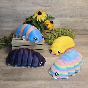 Könnte beinhalten: Vier farbenfrohe, segmentierte, insektenartige Figuren. Eine ist blau und rosa, eine andere gelb mit rosa Akzenten, eine dritte ist ein Regenbogen aus Farben und die letzte ist ein dunkles Lila mit Glitzer. Jede hat große, cartoonartige Augen und kleine Beine.
