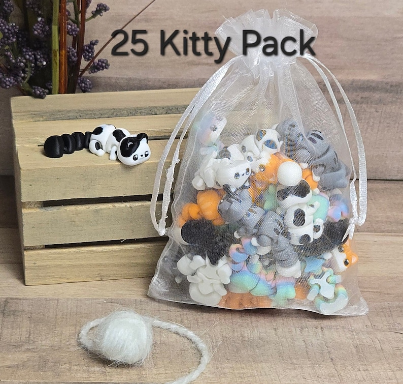K&ouml;nnte beinhalten: Ein wei&szlig;er Organzabag gef&uuml;llt mit 25 kleinen, bunten Katzenfiguren. Die Tasche ist mit einem wei&szlig;en Band gebunden und tr&auml;gt den Aufdruck "25 Kitty Pack".