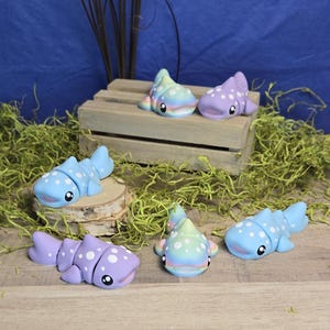 May include: A collection of handmade, whimsical whale shark figurines in various pastel colors. Each figurine features a unique design with white polka dots and expressive black eyes. Some are displayed on a wooden crate and a wood slice.