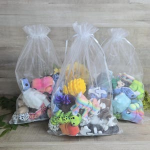 May include: Three sheer white organza drawstring bags filled with an assortment of small, colorful plastic animal figurines. The collection includes various creatures in shades of pink, yellow, green, blue, and white, featuring spotted and rainbow patterns.
