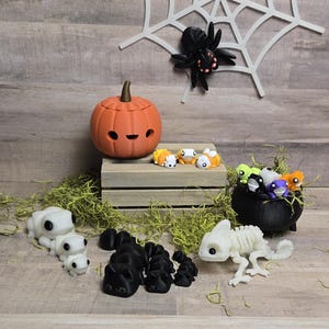 May include: A collection of Halloween decorations, including a small orange pumpkin with a smiling face, a black spider, and a variety of small white and colourful figurines. The decorations are arranged on a wooden shelf with a green moss-like material in the foreground.