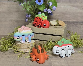 May include: Four colorful, handmade dinosaur figurines. One is red with green spikes, another is rainbow-colored, and a third is orange. The fourth is white with red spots and green spikes. They are displayed on a wooden surface with flowers.