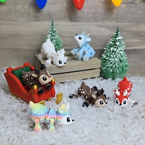May include: A set of six articulated reindeer figurines, including a red and white reindeer pulling a red sleigh, a white reindeer, a blue reindeer, a brown reindeer, and a rainbow reindeer. The figurines are on a white, fluffy surface with a wooden crate and a small pine tree in the background.