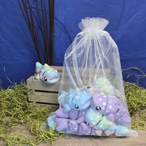 May include: A collection of colorful, plush toy axolotls in a sheer white drawstring bag. The axolotls are primarily blue, purple, and green with white polka dots. One axolotl sits outside the bag on a small wooden crate. The background is blue.