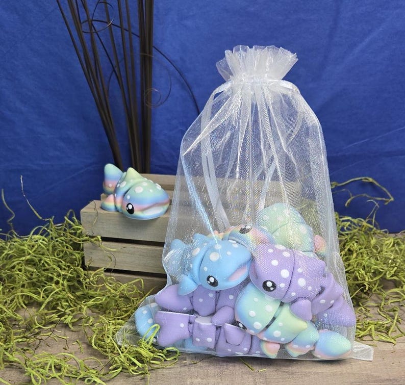 May include: A collection of colorful, plush toy axolotls in a sheer white drawstring bag. The axolotls are primarily blue, purple, and green with white polka dots. One axolotl sits outside the bag on a small wooden crate. The background is blue.