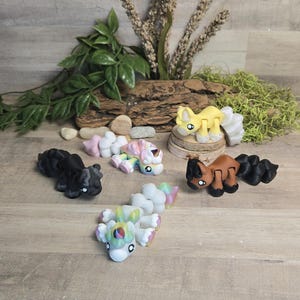 Puede incluir: Una colección de cinco figuras de animales articuladas con cuerpos segmentados. Las figuras incluyen una criatura negra sólida, una amarilla, una marrón con detalles negros y dos criaturas pastel multicolores con crines y colas de tonos arcoíris.