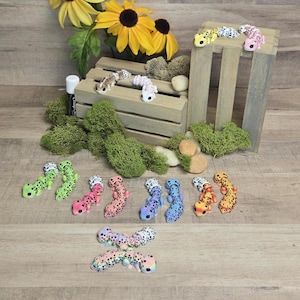May include: A collection of ten colourful gecko figurines, each with black spots. The geckos are arranged in a line on a light wood surface. The geckos are in various colours, including pink, blue, green, yellow, and orange.