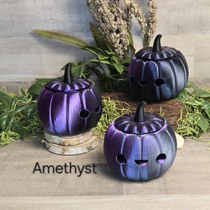May include: Three amethyst-colored pumpkin-shaped containers with black stems and smiling face cutouts. The containers are on a wooden surface with greenery in the background. The word "Amethyst" is printed on the surface.