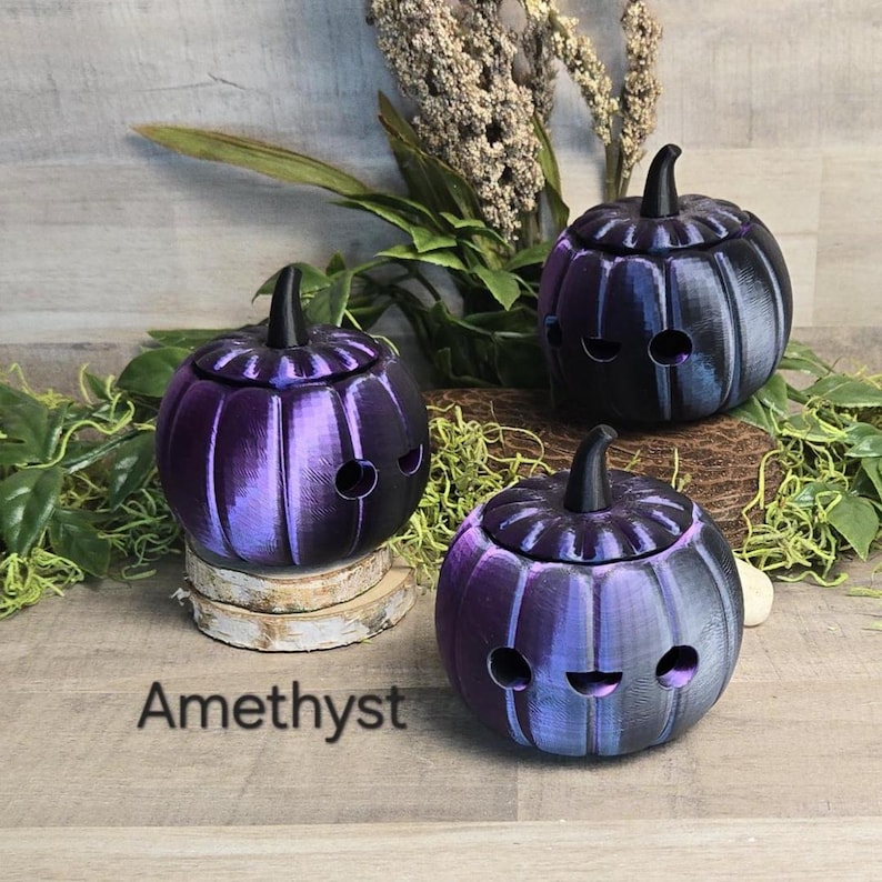 May include: Three amethyst-colored pumpkin-shaped containers with black stems and smiling face cutouts. The containers are on a wooden surface with greenery in the background. The word "Amethyst" is printed on the surface.
