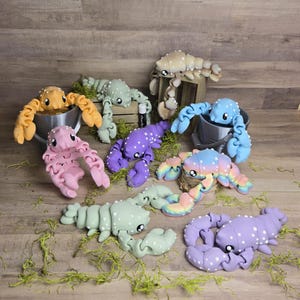 May include: Nine colourful crocheted lobster figurines in various poses. The lobsters are made of yarn and have a textured surface. They are displayed on a wooden surface with green moss.