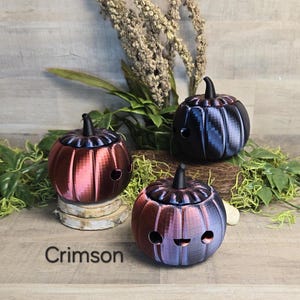 May include: Three decorative pumpkin-shaped containers in shades of crimson, black, and blue. Each pumpkin has a lid and a small, round opening on the side. The word "Crimson" is displayed at the bottom.