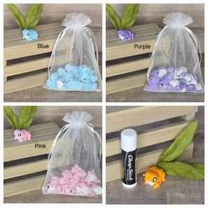 May include: Four small organza bags filled with miniature frog figurines. The bags are in blue, purple, pink, and white. A single orange frog figurine is on a wooden surface with a tube of ChapStick.