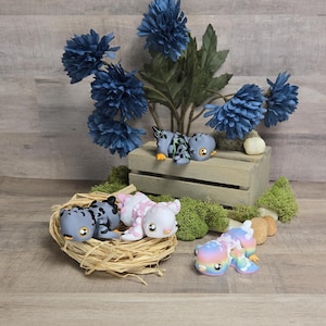 May include: Three small, hand-painted, ceramic figurines of stylised dragons. One dragon is grey and black, one is pink and white, and one is rainbow coloured. The dragons are posed in a playful, curled up position.