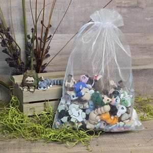 May include: A sheer white drawstring bag filled with small, colourful animal figurines. A few figurines are outside the bag, including a grey raccoon and a brown Bigfoot-like creature. The background includes a wooden crate and decorative greenery.