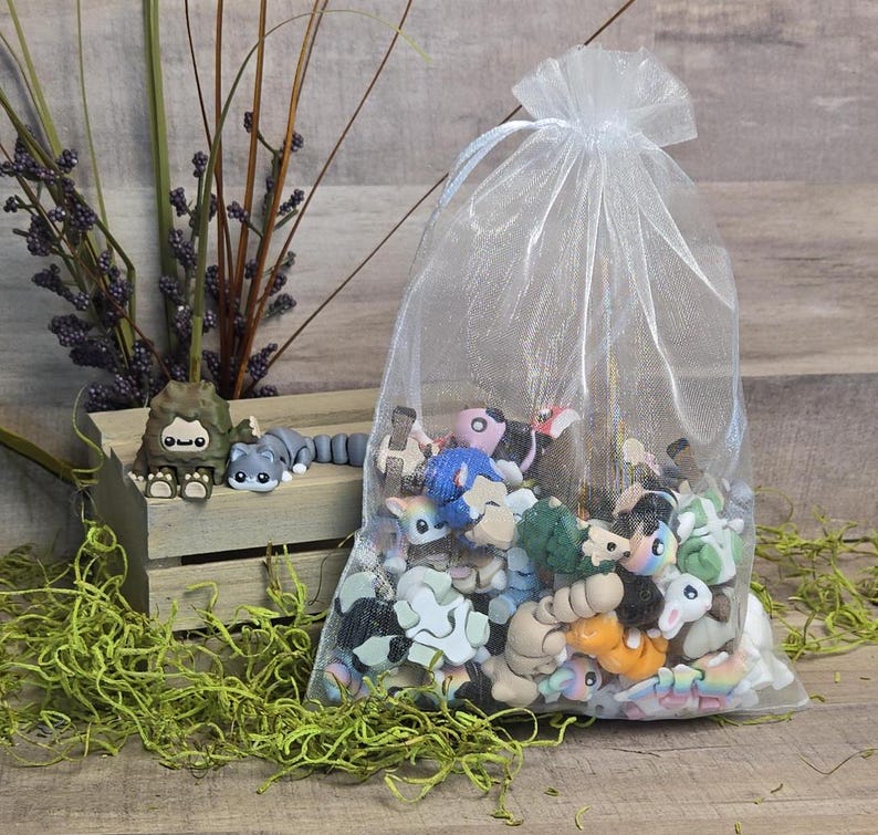 May include: A sheer white drawstring bag filled with small, colourful animal figurines. A few figurines are outside the bag, including a grey raccoon and a brown Bigfoot-like creature. The background includes a wooden crate and decorative greenery.