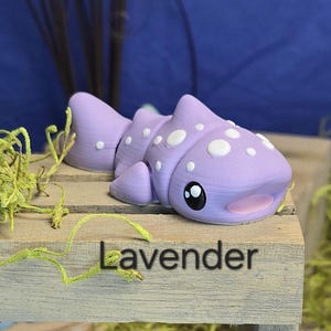 May include: A lavender-colored, segmented, cartoon-style fish figurine with white polka dots and large black eyes. The word "Lavender" is printed in black on the wooden surface below the figurine. The figurine is displayed on a wooden surface with green moss.