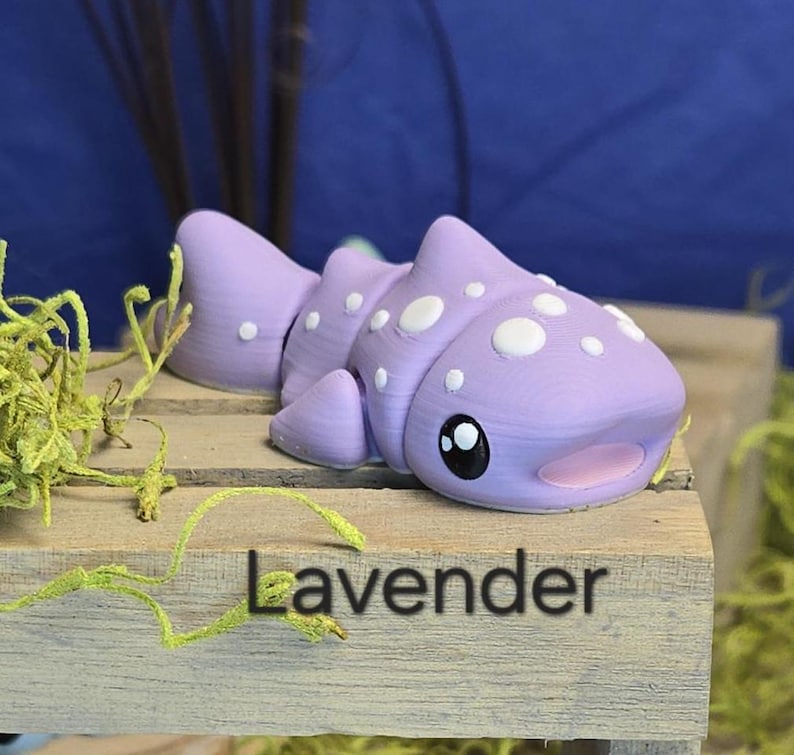 May include: A lavender-colored, segmented, cartoon-style fish figurine with white polka dots and large black eyes. The word "Lavender" is printed in black on the wooden surface below the figurine. The figurine is displayed on a wooden surface with green moss.