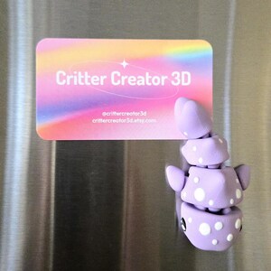 May include: A lavender 3D-printed creature magnet with white polka dots and black eyes. The magnet is next to a business card with the text "Critter Creator 3D" and social media information.