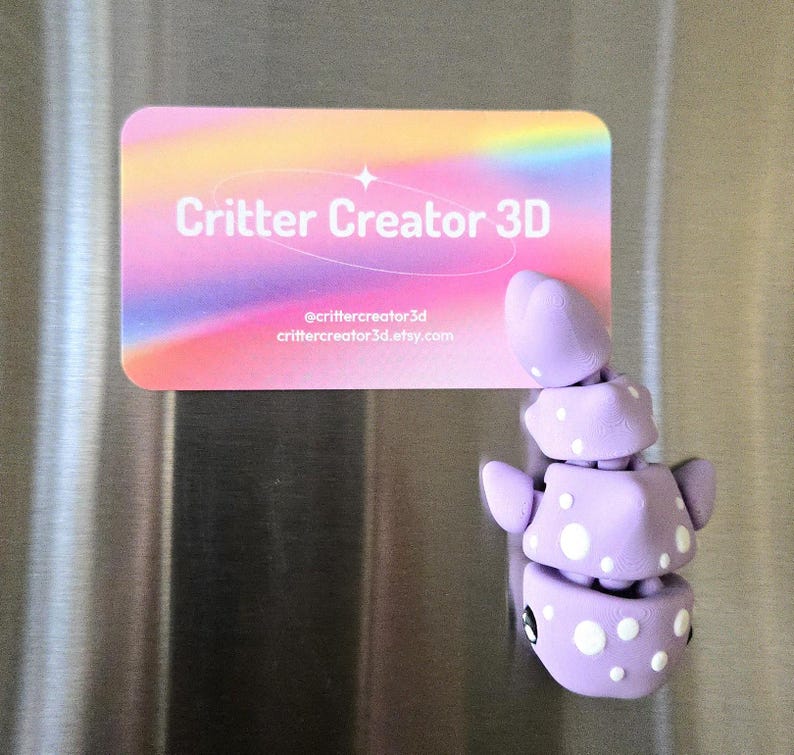 May include: A lavender 3D-printed creature magnet with white polka dots and black eyes. The magnet is next to a business card with the text "Critter Creator 3D" and social media information.