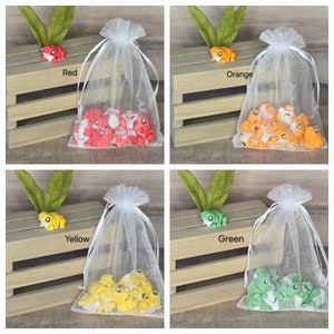 May include: Four white organza bags filled with small frog figurines. Each bag has a different color frog: red, orange, yellow, and green. The bags are sitting on a wooden crate.