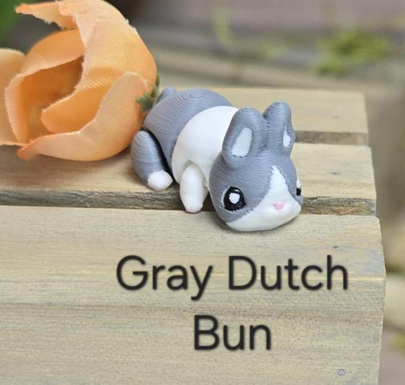 K&ouml;nnte beinhalten: Eine grau, wei&szlig; und schwarze Hasenfigur mit gro&szlig;en Ohren und einer rosa Nase. Die Figur steht auf einer Holzoberfl&auml;che mit dem Text "Gray Dutch Bun" in Schwarz gedruckt. Im Hintergrund befindet sich eine orangefarbene Blume.