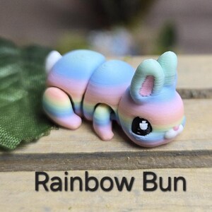 K&ouml;nnte beinhalten: Eine farbenfrohe, regenbogenfarbene Hasenfigur mit einem segmentierten K&ouml;rper und gro&szlig;en Ohren. Der Hase liegt auf einer Holzoberfl&auml;che, darunter der Text "Rainbow Bun". Die Farben des Hasen sind Rosa, Blau und Gelb.