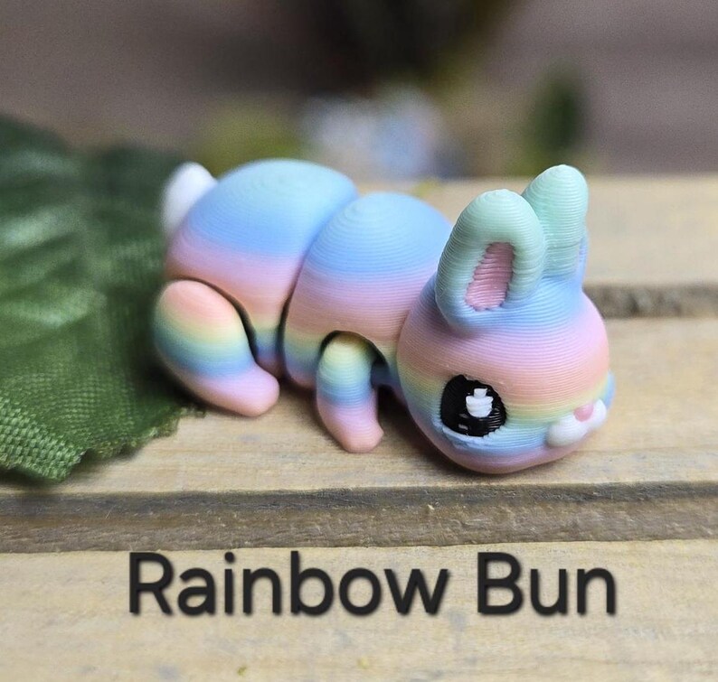 K&ouml;nnte beinhalten: Eine farbenfrohe, regenbogenfarbene Hasenfigur mit einem segmentierten K&ouml;rper und gro&szlig;en Ohren. Der Hase liegt auf einer Holzoberfl&auml;che, darunter der Text "Rainbow Bun". Die Farben des Hasen sind Rosa, Blau und Gelb.