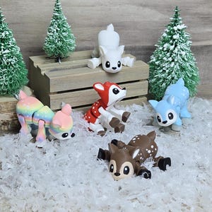 May include: Five small, articulated, deer-like figurines in various colors, including white, red, blue, rainbow, and brown, are posed on a white, fluffy surface. The figurines are made of plastic and have a whimsical, cartoonish style.