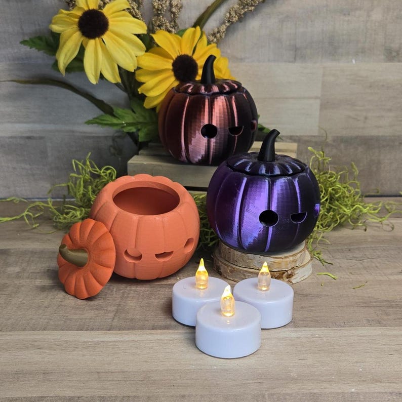 May include: A collection of Halloween-themed pumpkin decorations. The image features orange, purple, and dark red pumpkin-shaped containers with carved faces. Battery-operated tea lights are placed in front of the pumpkins, with sunflowers in the background.
