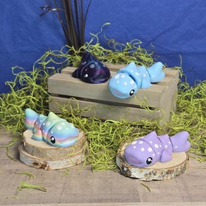 May include: Four colorful whale shark figurines. One is dark blue with white spots, another is light blue with white spots, a third is pastel rainbow with white spots, and the last is purple with white spots. Each figurine is on a wooden slice.