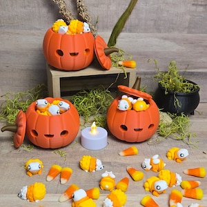 Puede incluir: Tres contenedores naranja en forma de calabaza con tapas, llenos de pequeños dulces blancos y naranjas. Los contenedores están sobre una superficie de madera con una variedad de pequeños dulces blancos y naranjas esparcidos a su alrededor.