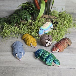 May include: Six articulated armadillo figurines in various colors including pastel rainbow, solid yellow, brown, grey, iridescent green, and reddish-brown. Each small animal figure features a segmented shell design and a light-colored snout.