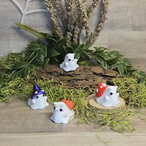 Peut inclure: Plusieurs figurines de fantômes blancs, chacune avec des accessoires uniques. L'une porte un chapeau de sorcière violet, une autre un chapeau de champignon rouge et une troisième une citrouille orange. Les figurines sont sur une bûche en bois avec de la mousse verte.