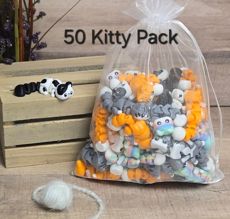 K&ouml;nnte beinhalten: Ein wei&szlig;er Netzbeutel gef&uuml;llt mit 50 kleinen, bunten Katzen-Anh&auml;ngern. Der Beutel ist mit "50 Kitty Pack" beschriftet.