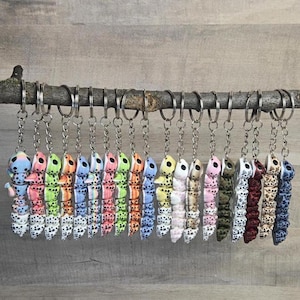 Puede incluir: Una colección de llaveros de oruga coloridos colgando de una rama de madera. Cada llavero presenta una combinación de colores única, incluyendo rosa, azul, verde, naranja y marrón, con lunares negros. Los llaveros están unidos a anillos de metal plateado.