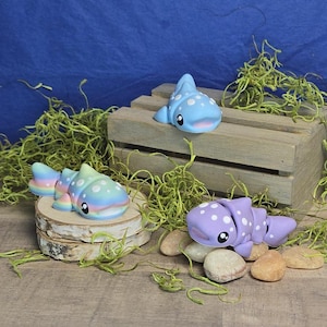 May include: Three whimsical, handmade clay whale shark figurines in shades of blue, pastel rainbow, and lavender. Each whale shark has white polka dots and a smiling face. One rests on a wooden crate, another on a wood slice, and the third on small stones.