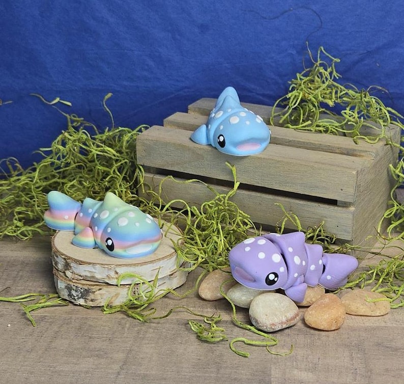 May include: Three whimsical, handmade clay whale shark figurines in shades of blue, pastel rainbow, and lavender. Each whale shark has white polka dots and a smiling face. One rests on a wooden crate, another on a wood slice, and the third on small stones.